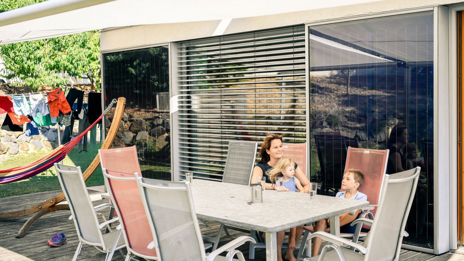 Eine Frau und zwei Kinder sitzen auf der Terrasse vor dem Haus. 