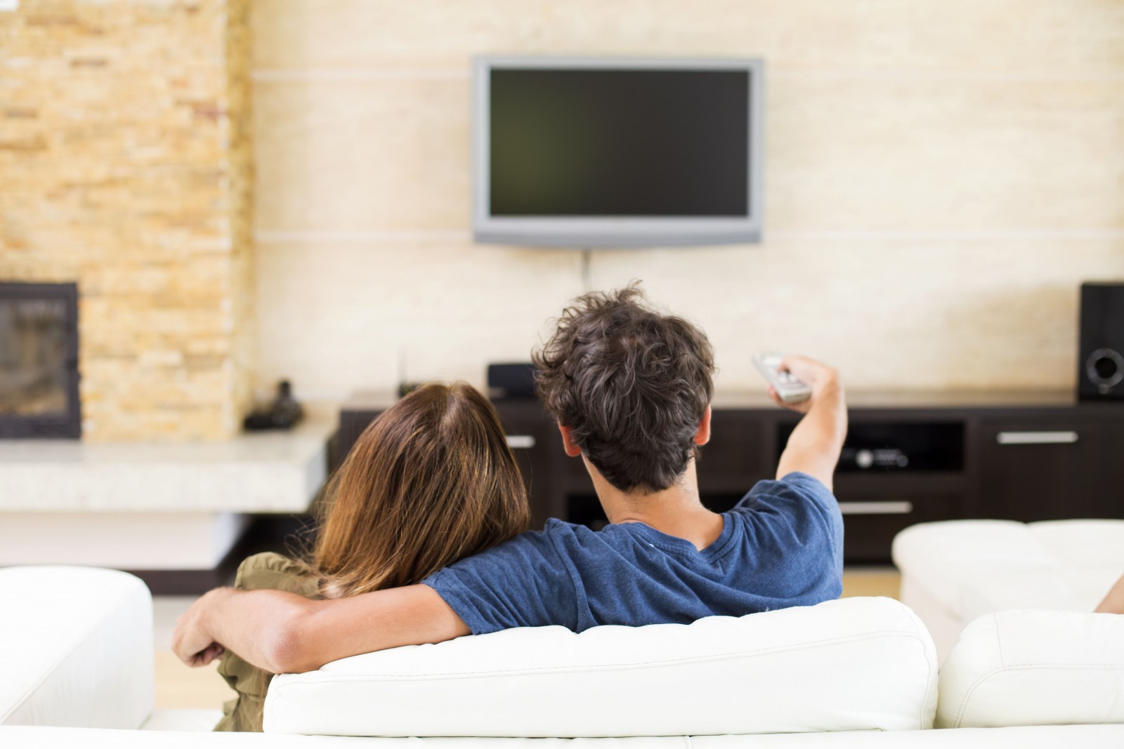 Ein junges Paar sitzt im Wohnzimmer vor dem Fernseher. Der Mann hält die Fernbedienung zum EInschalten des Gerätes in der Hand.