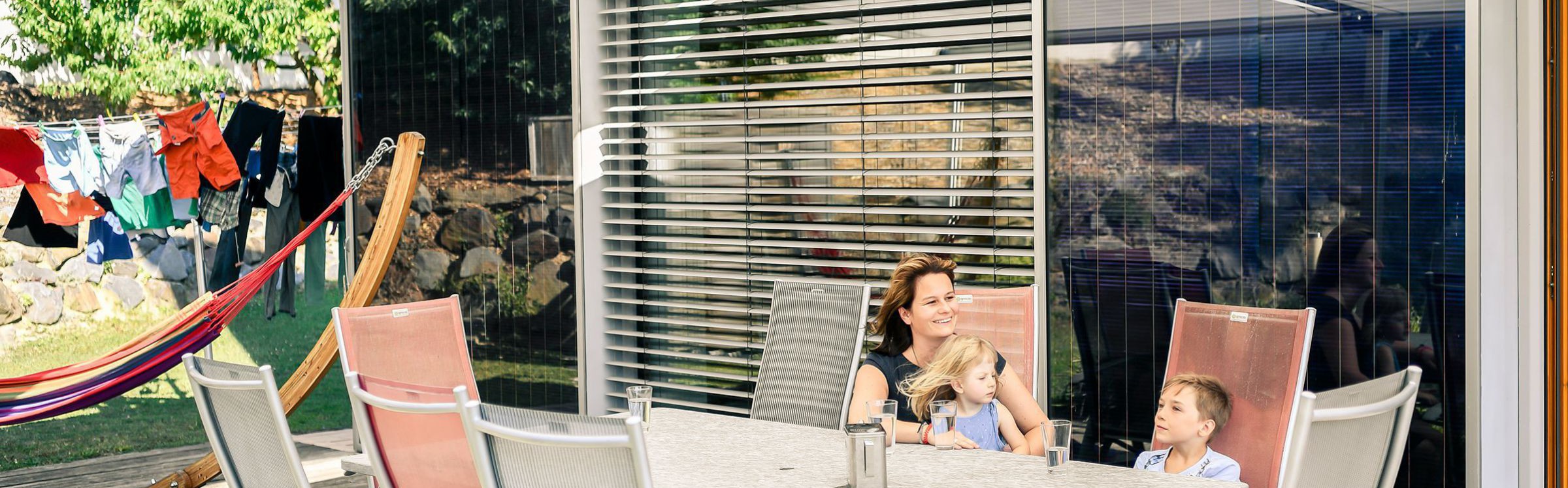 Eine Frau und zwei Kinder sitzen auf der Terrasse vor dem Haus.