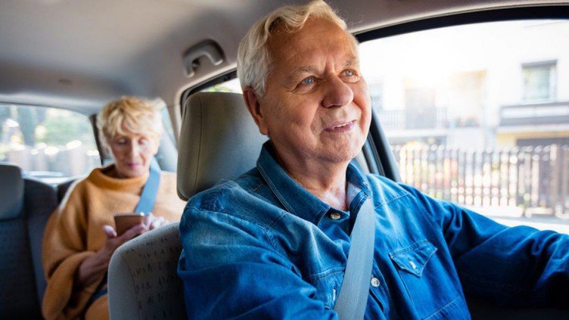 Älterer Herr fährt Auto und ältere Dame sitzt am Rücksitz mit Handy